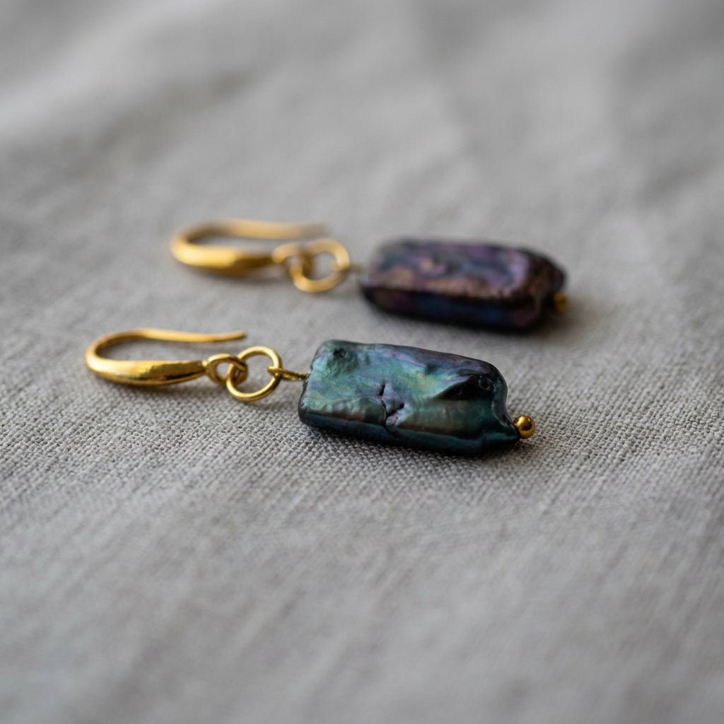 Dark blue square baroque pearl drop earrings lying side by side on pale linen, showing green-blue and purple iridescence with gold-filled hooks and small gold bead tips.