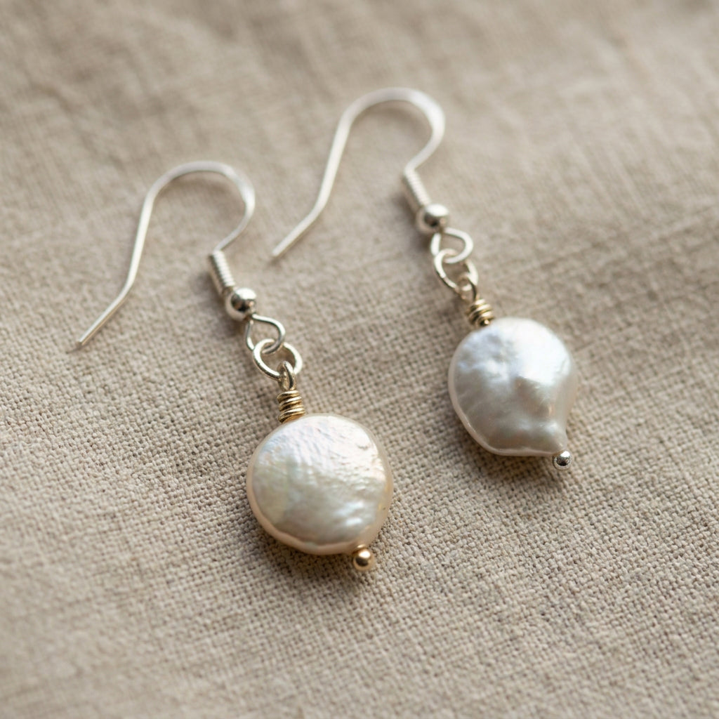 Pair of white coin pearl drop earrings on beige linen, showing softly textured nacre surfaces, sterling silver hooks and connecting links.