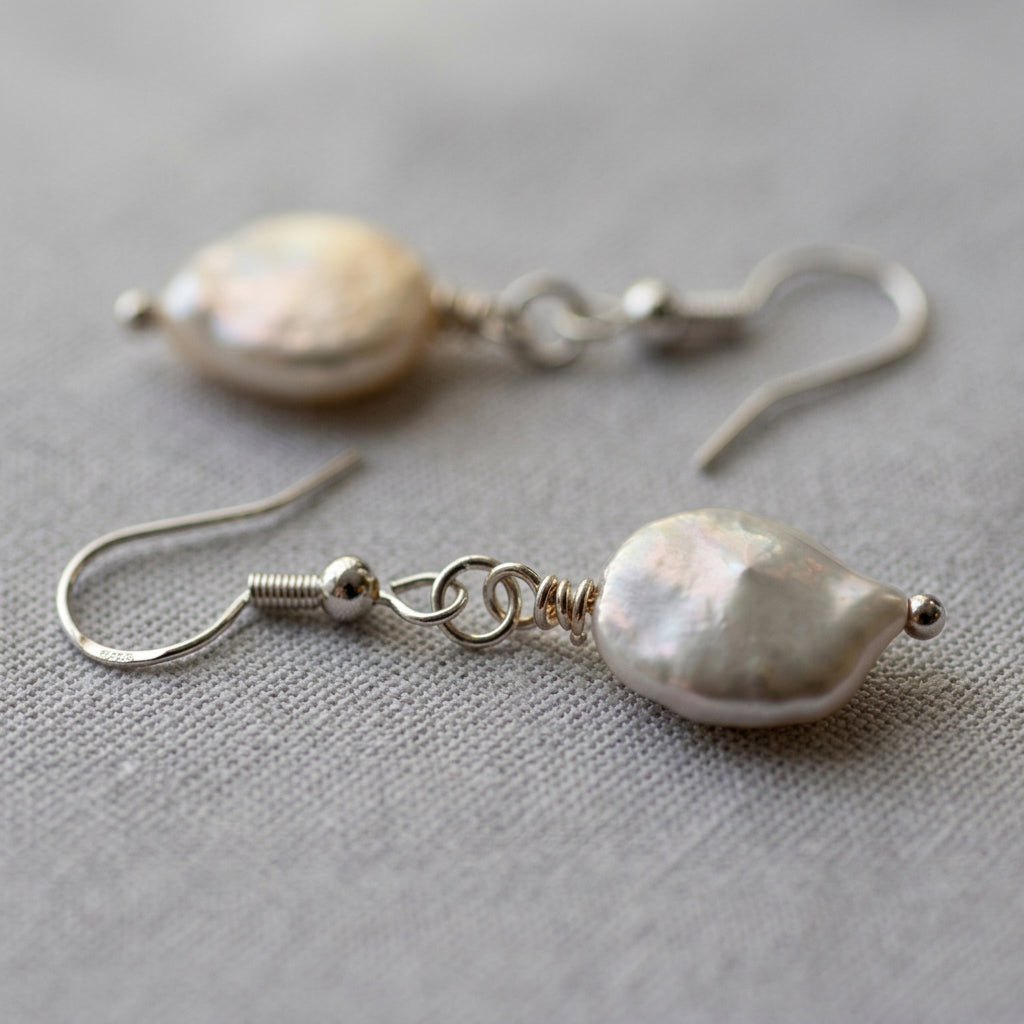 Close-up of white coin pearl drop earrings on grey linen, showing softly iridescent surfaces, sterling silver hooks, connecting rings and small silver bead tips.