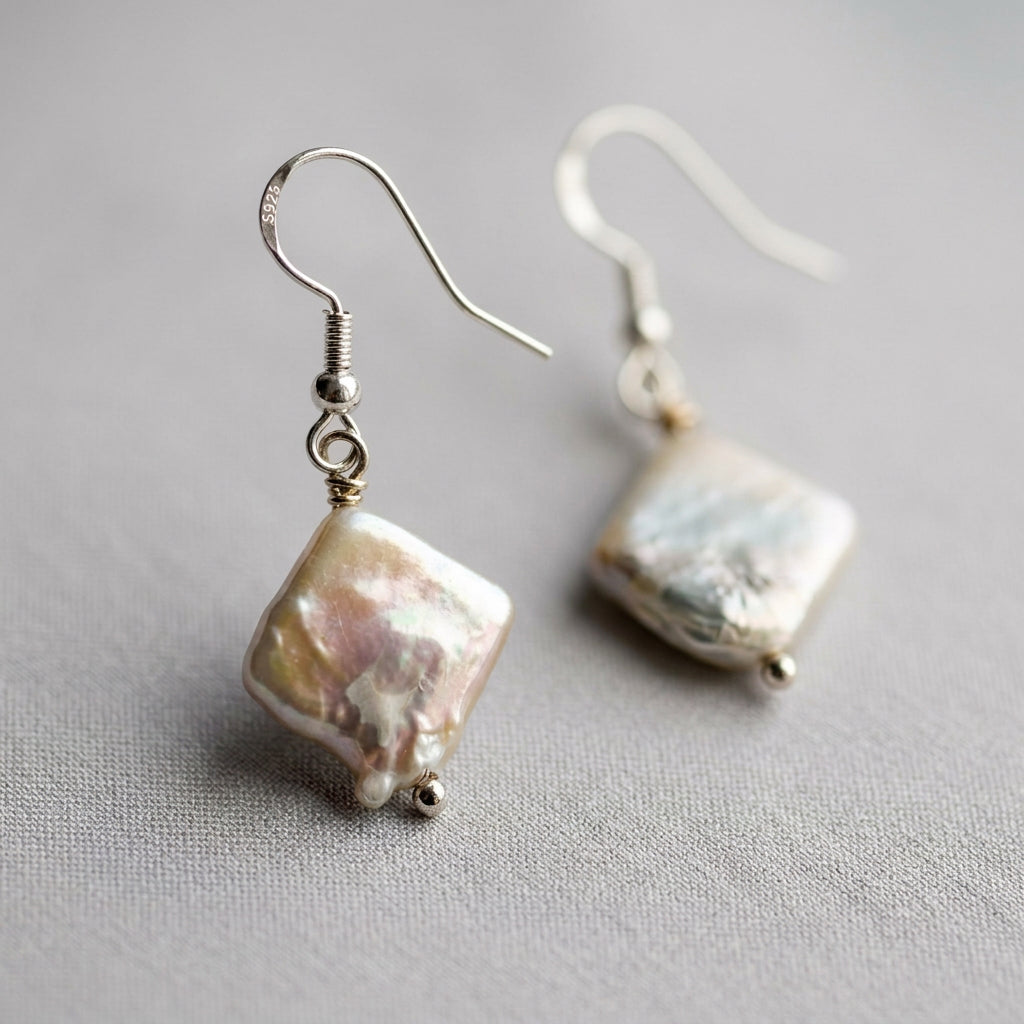 Close-up of white square baroque pearl drop earrings on pale linen, showing textured pink-gold iridescence and sterling silver hooks, one marked 925.