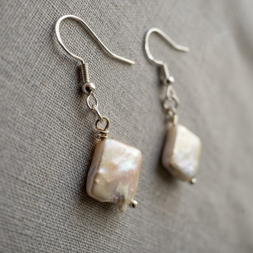 White square baroque pearl drop earrings lying side by side on grey linen, showing soft pink-gold iridescence, sterling silver hooks and connecting links.