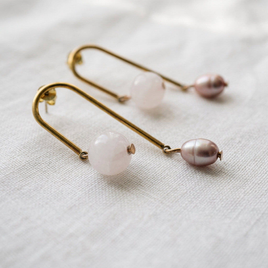 Pair of gold-coated arch drop earrings resting on white linen, each with a pale pink quartz bead on the shorter side and a lustrous mauve-pink baroque pearl on the longer side.