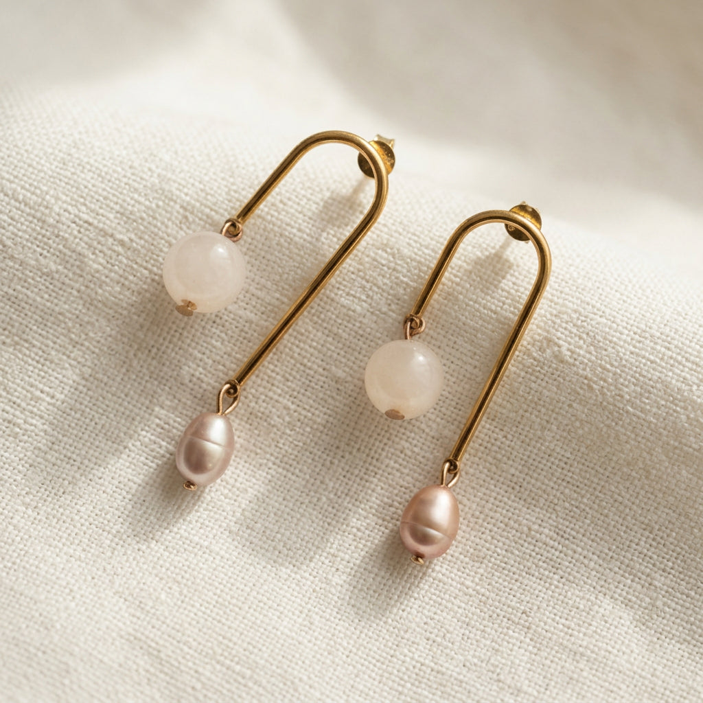 Pair of gold-coated arch drop earrings arranged side by side on cream linen, with pale pink quartz beads on the shorter arms and pink baroque pearls on the longer arms.