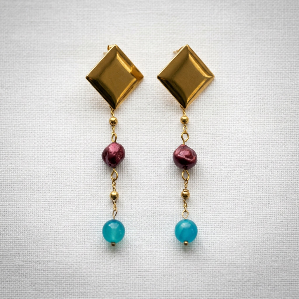 Pair of statement drop earrings arranged side by side on white linen, with polished diamond-shaped gold-coated studs, burgundy baroque pearls and bright blue topaz beads linked by fine beaded chains.