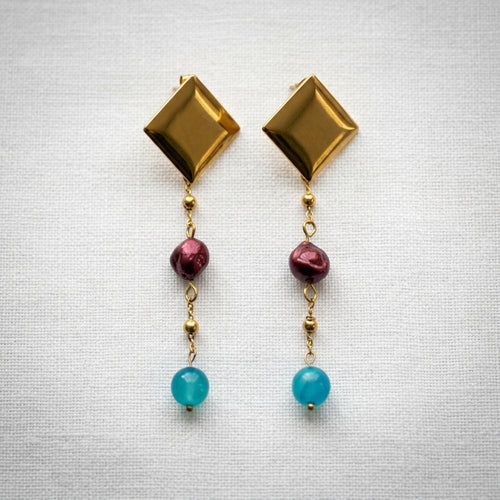 Pair of statement drop earrings arranged side by side on white linen, with polished diamond-shaped gold-coated studs, burgundy baroque pearls and bright blue topaz beads linked by fine beaded chains.