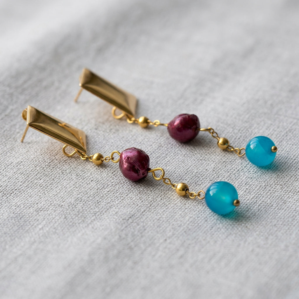 Angled close-up of burgundy baroque pearl and bright blue topaz drop earrings on grey linen, showing the gold-coated pyramid studs, exposed posts and fine chains with small gold bead accents.