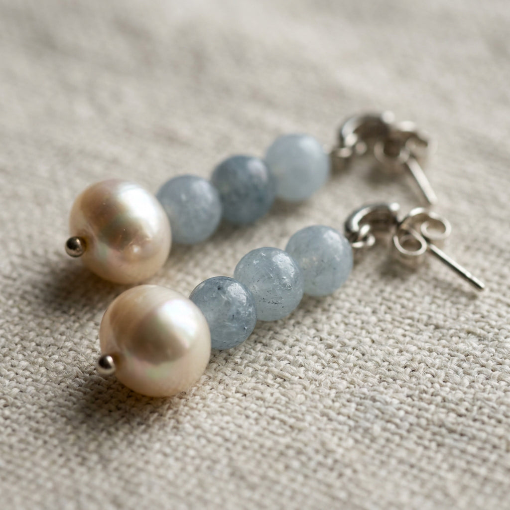 Close-up of aquamarine and baroque pearl drop earrings on textured cream linen, showing natural inclusions in the pale blue beads, creamy pearl lustre and sterling silver butterfly backs.⠋