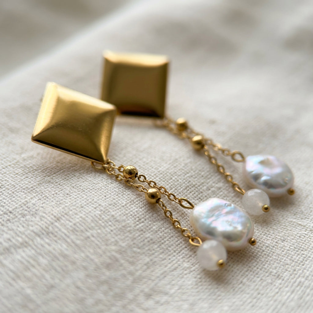 Pair of gold-coated drop earrings on cream linen, with polished square studs and twin beaded chains ending separately in iridescent oval white baroque pearls and pale pink quartz beads.