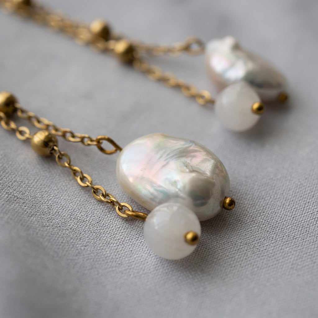 Close-up of an oval white baroque pearl with pink-green iridescence beside a pale pink quartz bead, suspended from separate gold-coated beaded chains on grey fabric.