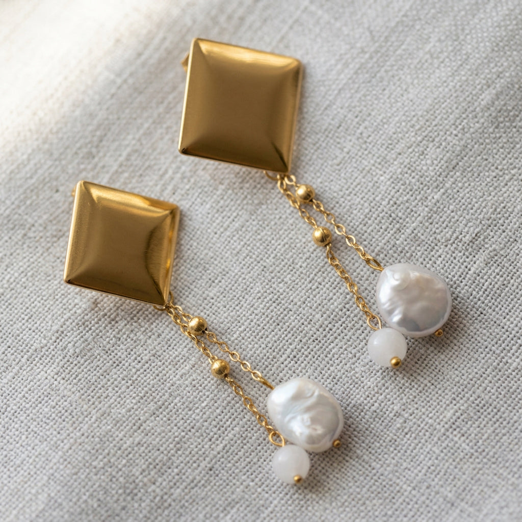 Pair of gold-coated drop earrings arranged diagonally on grey linen, with polished square studs and twin beaded chains ending separately in oval white baroque pearls and pale pink quartz beads.