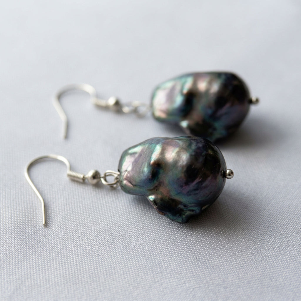 Low-angle close-up of dark keshi pearl earrings on white fabric, highlighting their folded surfaces, silvery-blue lustre and sterling silver hooks.