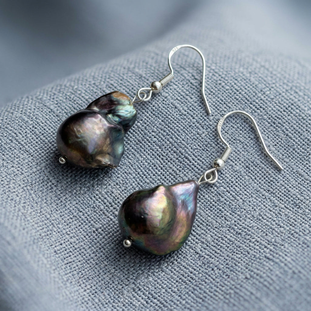 Close-up of a dark blue keshi pearl earring worn on a sterling silver hook, showing its uneven contours and blue-purple iridescent highlights.