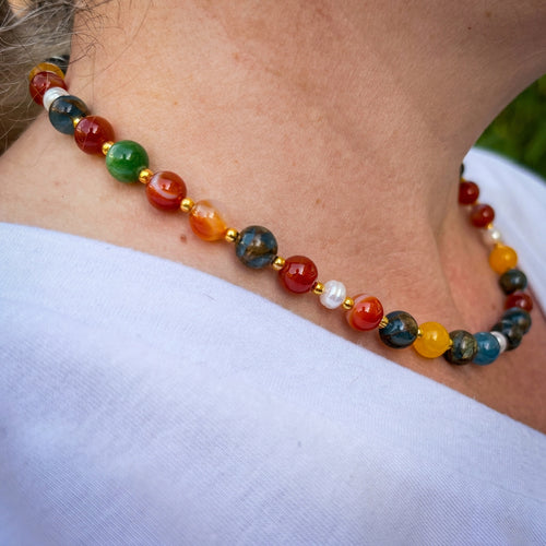 Side view of a colourful beaded necklace worn at the collarbone, showing blue, orange-red, yellow and green stones, white baroque pearls and gold-coloured spacers.