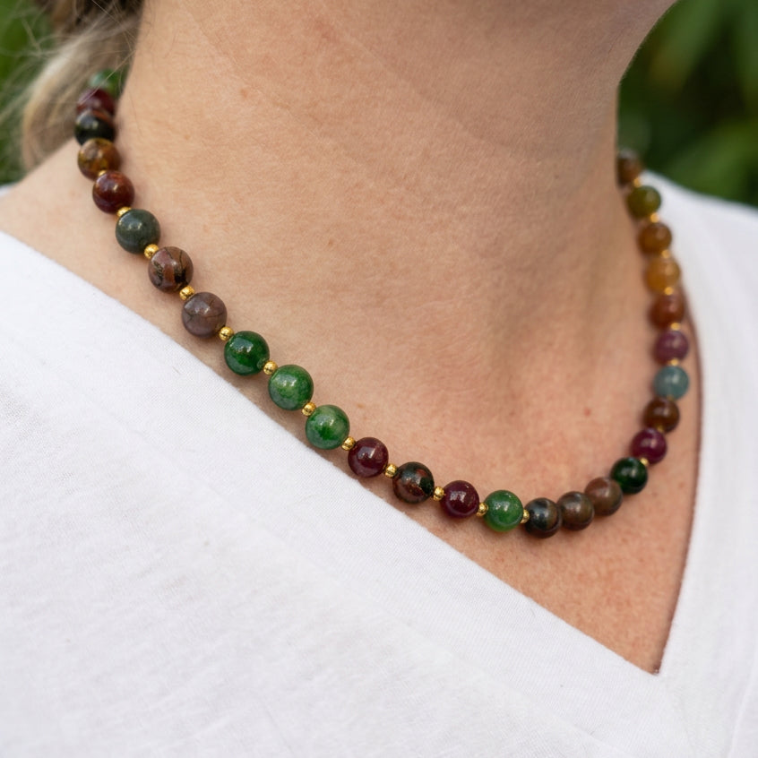 Side view of a jasper, tourmaline, jade and garnet necklace worn above a white V-neck top, showing green, burgundy and earthy brown beads with gold-coloured spacers.