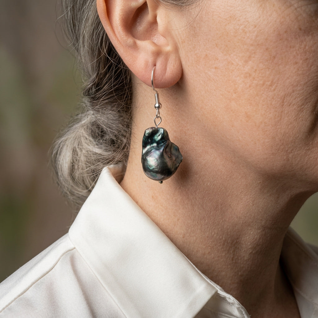 Large dark blue keshi pearl drop earring on a sterling silver hook, worn above a white shirt collar, showing its irregular sculptural shape and silvery green-blue lustre.
