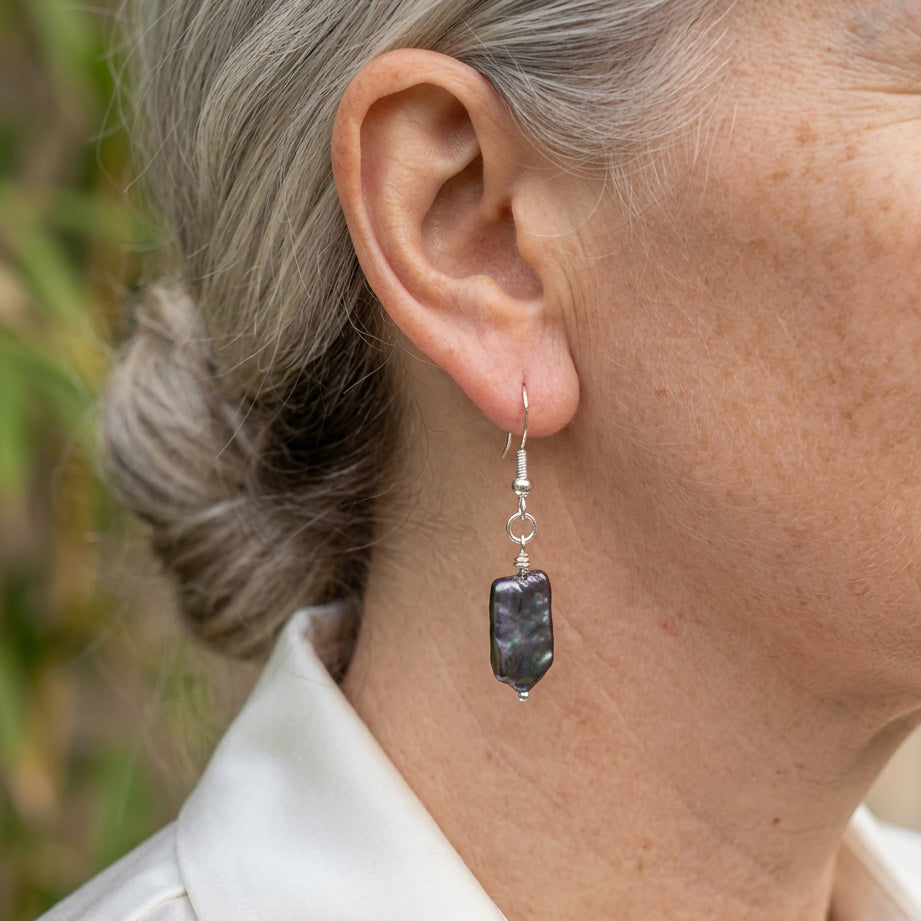 Dark blue square baroque pearl drop earring worn on an ear, shown side-on with subtle green-purple iridescence and a sterling silver hook.