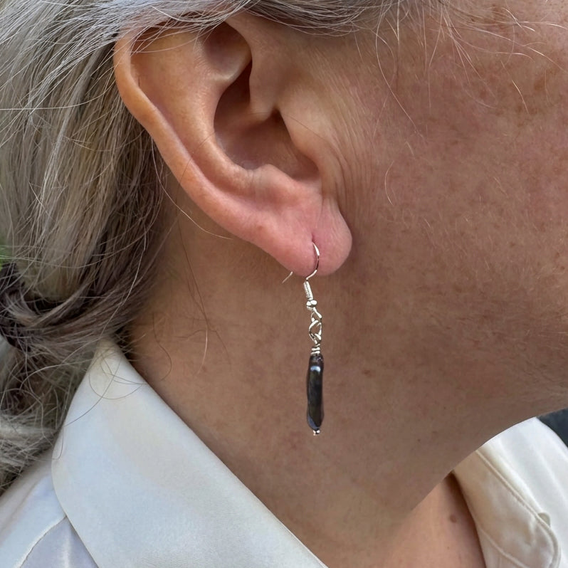Side-on close-up of a dark blue square baroque pearl drop earring worn on an ear, showing blue-purple highlights, sterling silver wire wrapping and connecting rings.