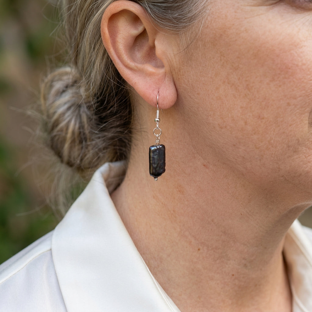 Close-up of a dark blue square baroque pearl drop earring worn on an ear, showing its textured surface, sterling silver hook and connecting rings.