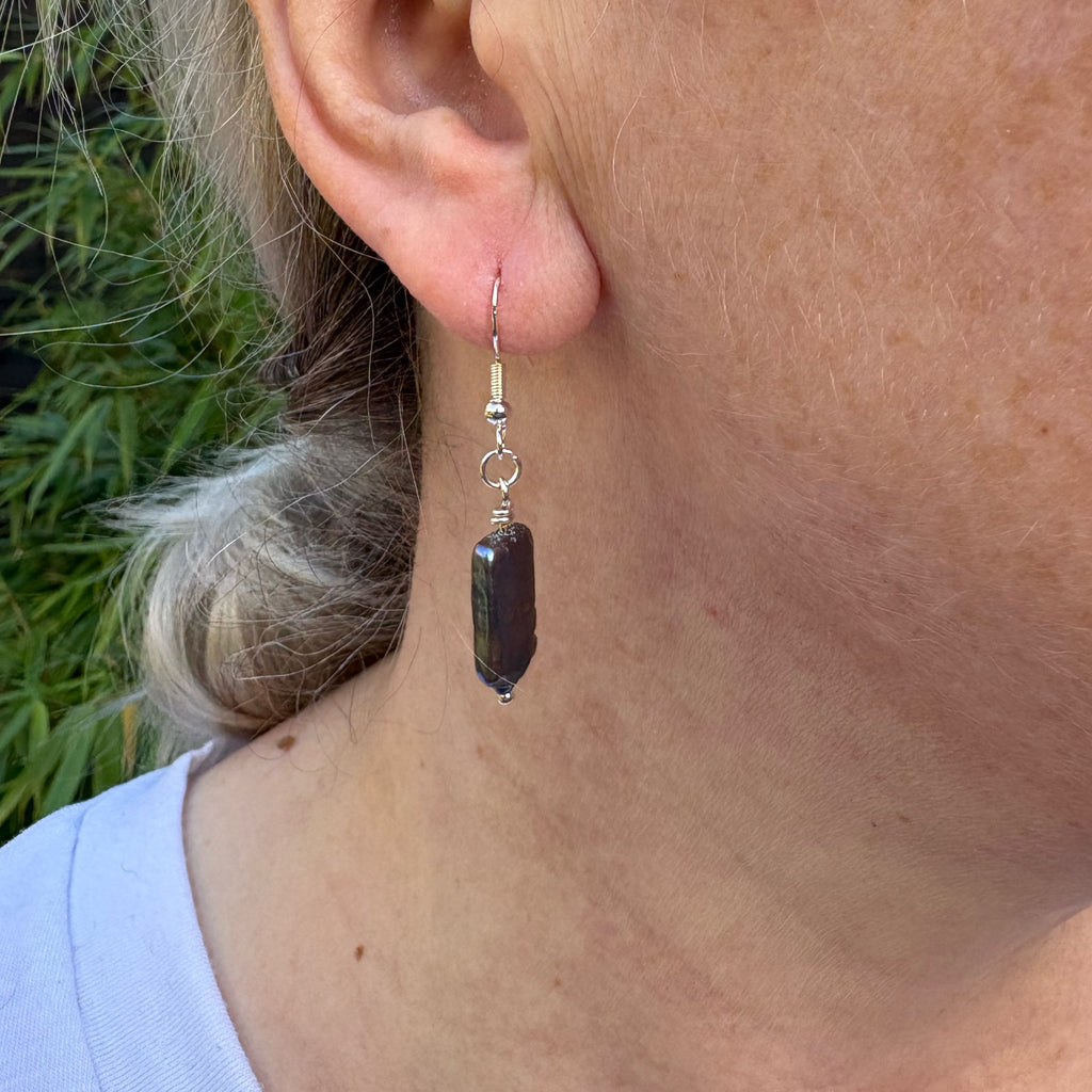 Side view of a dark blue square baroque pearl drop earring worn below the earlobe, showing its slim profile and sterling silver hook.