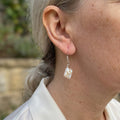White square baroque pearl drop earring worn on an ear, showing its textured iridescent surface, sterling silver hook and connecting rings.
