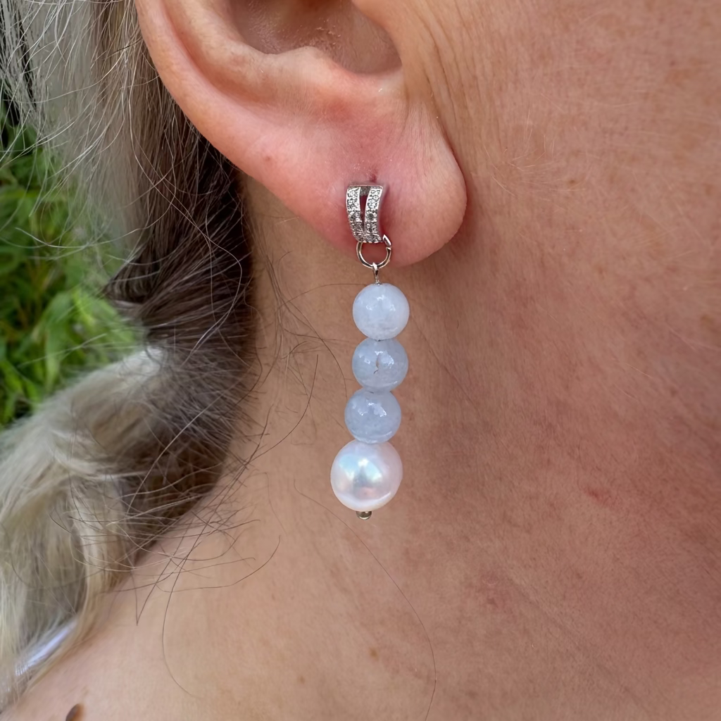 Close-up of an aquamarine and baroque pearl drop earring worn on an ear, with three pale blue beads above a lustrous white pearl and a crystal-set sterling silver stud.⠋