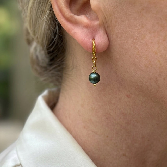 Close-up of a peacock pearl drop earring worn above a white shirt collar, showing its deep green lustre, gold-filled links and textured hook.