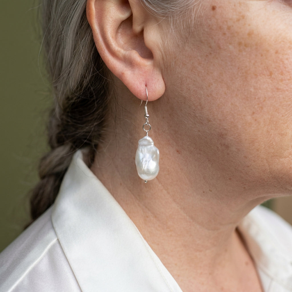 White Keshi pearl drop earring worn below the earlobe, showing its elongated irregular shape, lustrous rippled surface and sterling silver hook with connecting rings.