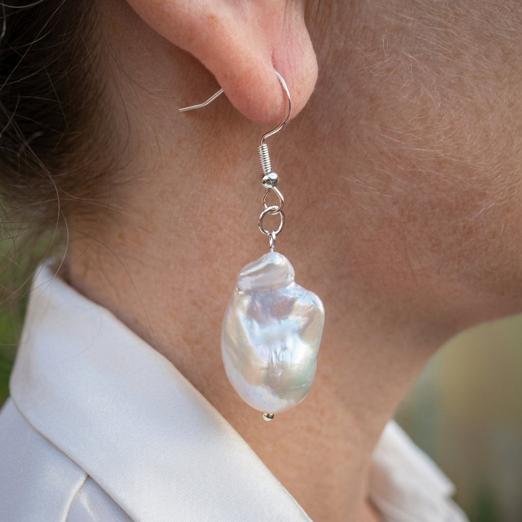 White Keshi pearl drop earring worn on an ear, showing its elongated irregular shape, rippled iridescent surface and sterling silver hook with connecting rings.