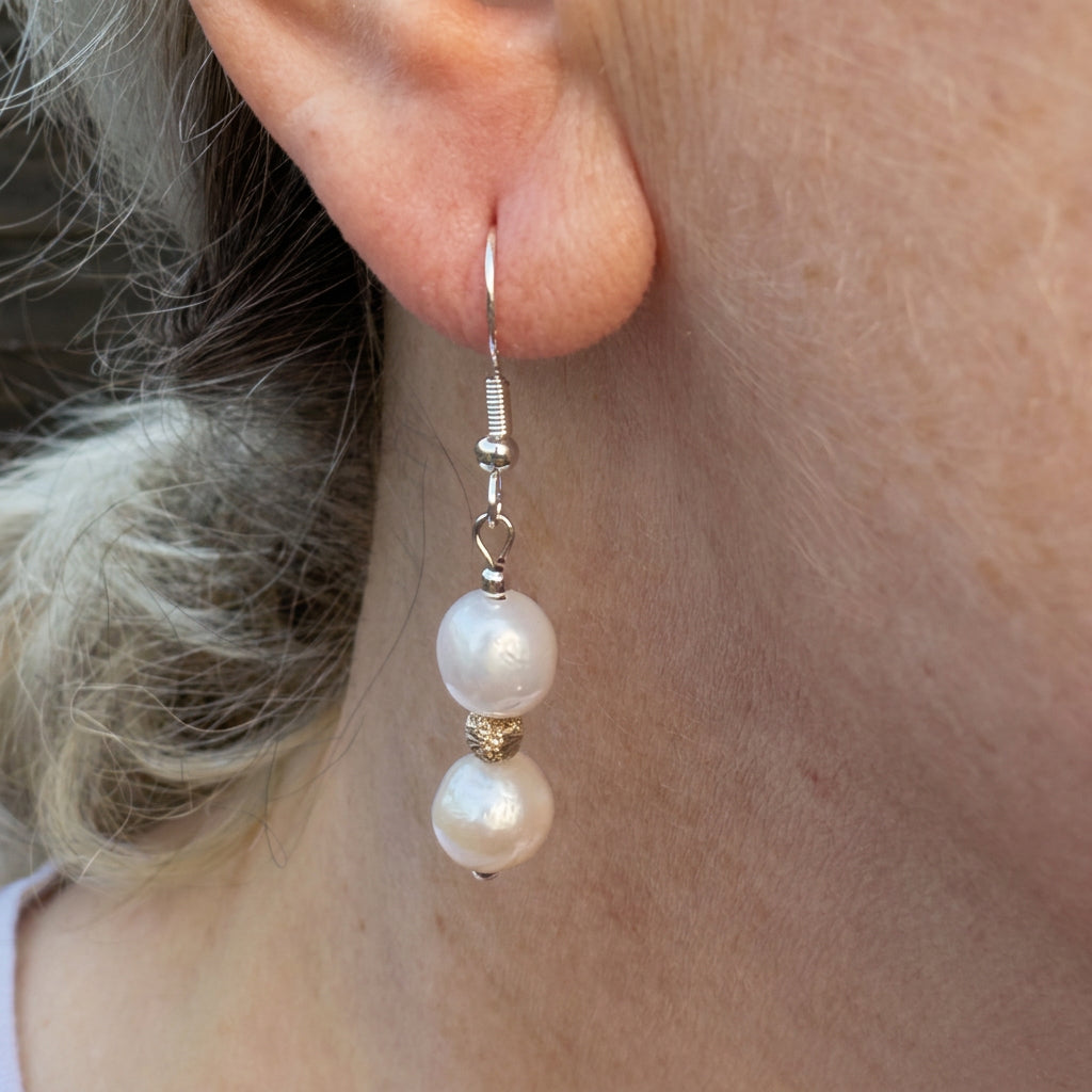 Close-up of a baroque pearl drop earring worn on an ear, with two creamy white pearls separated by a textured gold-coloured spacer on a sterling silver shepherd hook.