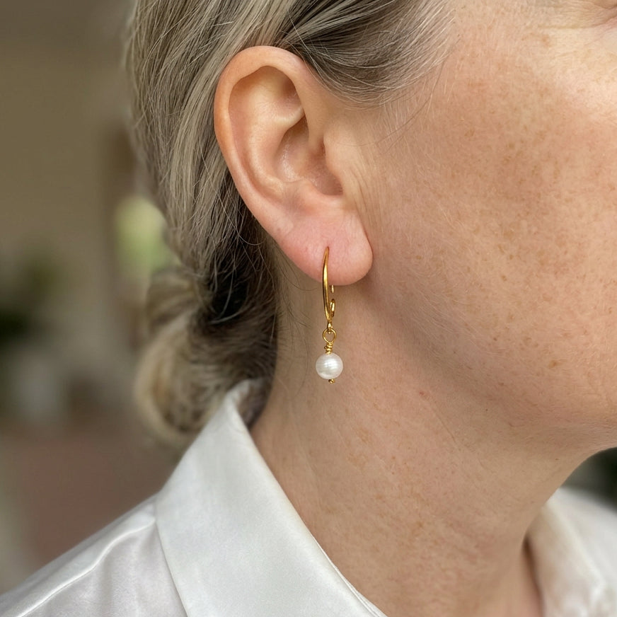 White baroque pearl drop earring worn on an ear, with a small lustrous pearl suspended from a curved gold-filled hook and wire-wrapped link.