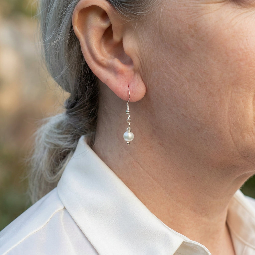 White baroque pearl drop earring worn on an ear, with a small lustrous pearl suspended from a sterling silver hook and connecting links.