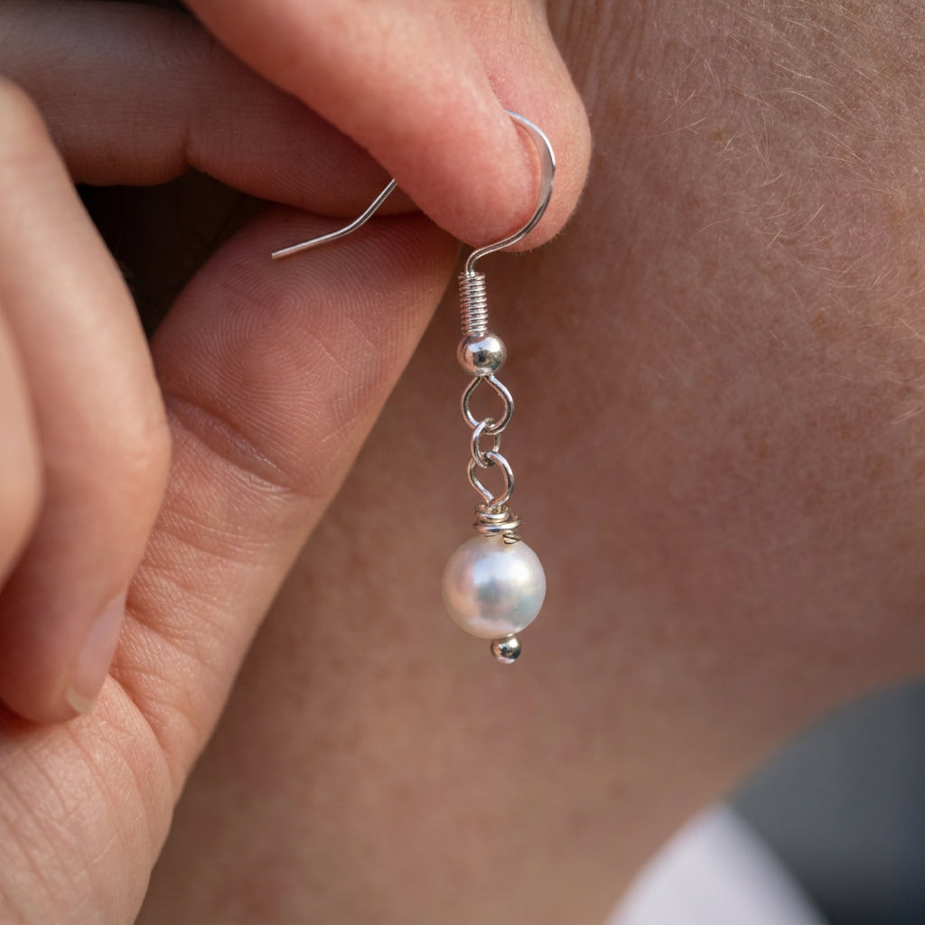 White baroque pearl drop earring held beside an ear, showing the lustrous pearl, sterling silver hook, connecting links and small silver bead tip.
