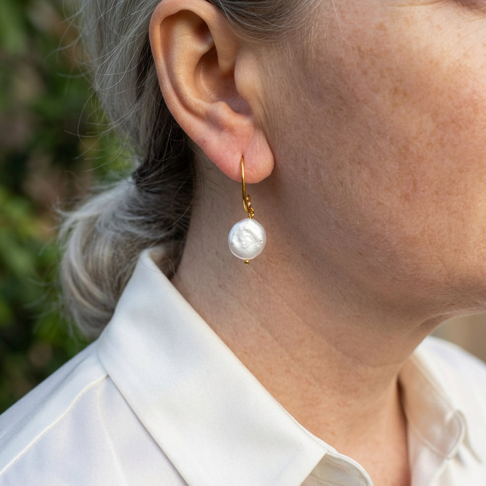 White coin pearl drop earring worn on an ear, showing its softly iridescent face, curved gold-filled hook and small gold bead tip.