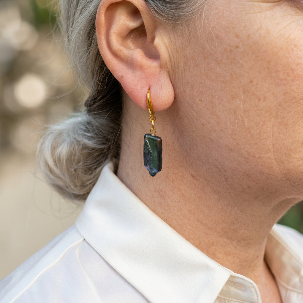 Dark blue rectangular baroque pearl drop earring on a gold-filled hook, worn above a white shirt collar, showing its irregular edges and deep green-blue iridescent surface.