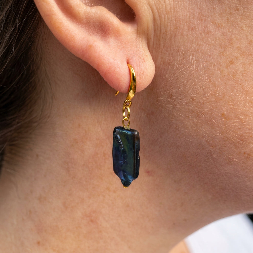 Close-up of a dark blue square baroque pearl drop earring worn on an ear, showing green-blue iridescence, a gold-filled hook and a twisted connecting link.