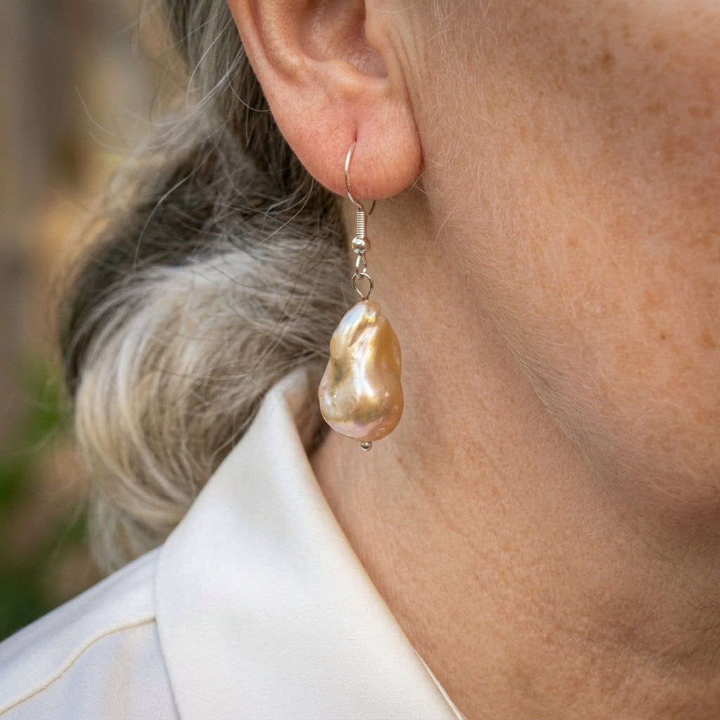 Cream keshi pearl drop earring worn on an ear, showing a large irregular pearl with golden-peach tones and pink-green iridescence suspended from a sterling silver hook.