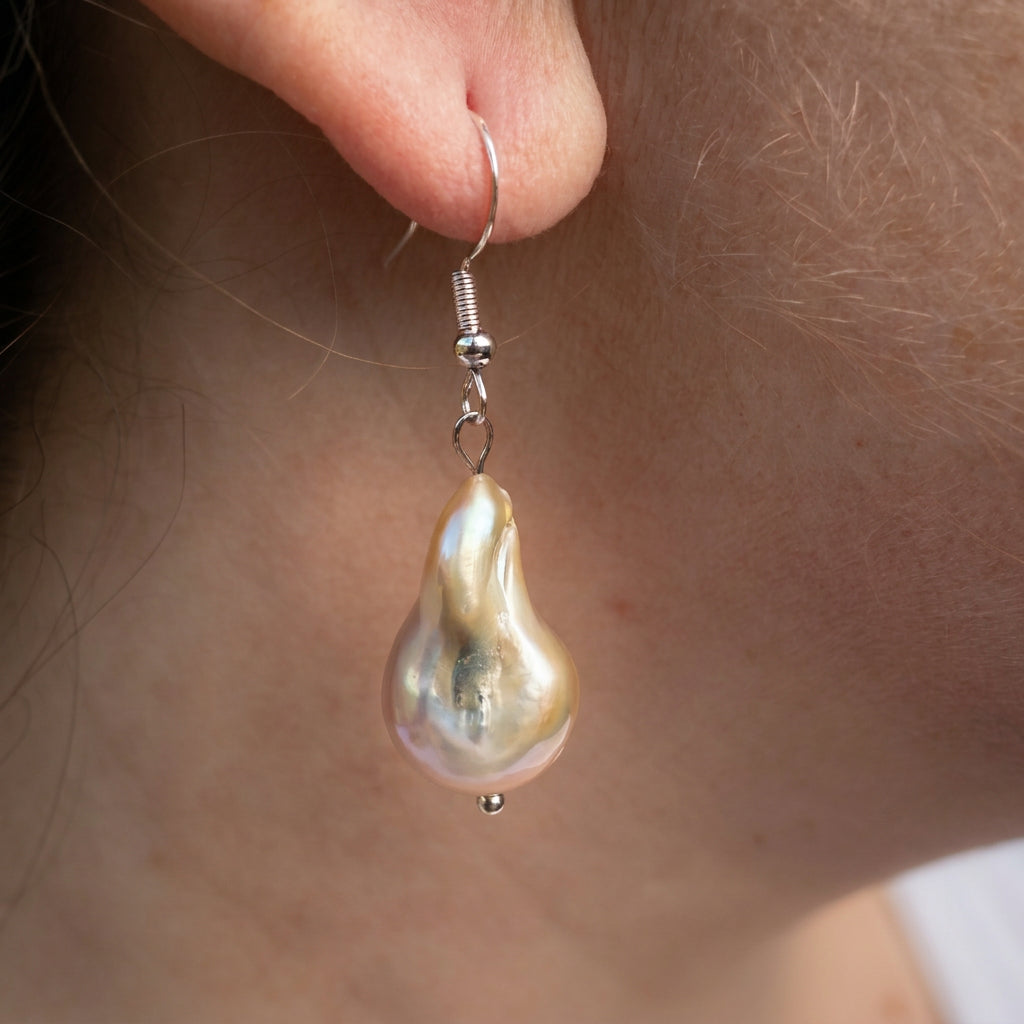 Side view of a cream keshi pearl earring worn on an ear, showing an elongated irregular pearl with golden, pink and green reflections suspended from a sterling silver hook.