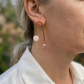 Gold-plated arch earring with a pale pink quartz bead on the shorter arm and a blush baroque pearl on the longer arm, worn above a white shirt collar.