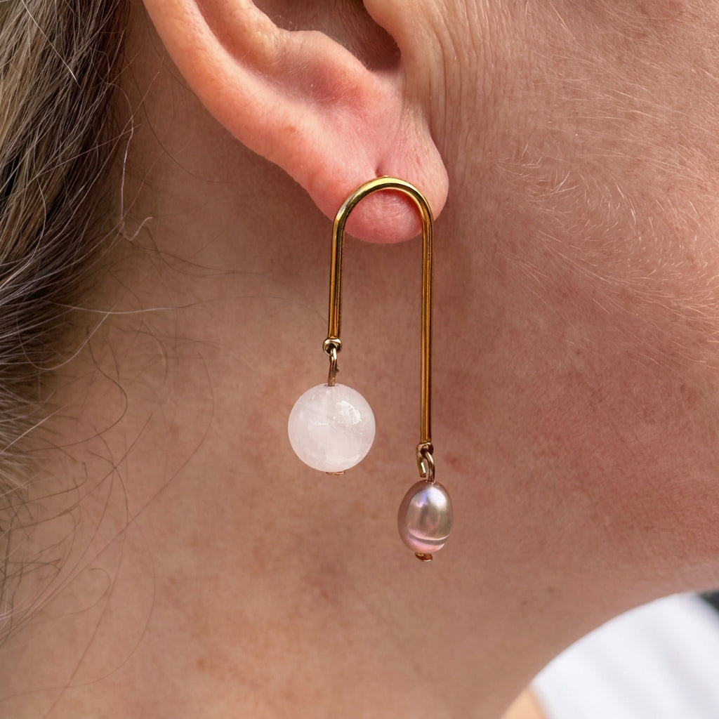 Side view of a gold-coated arch drop earring worn on an ear, with a pale pink quartz bead and a smaller mauve-pink baroque pearl hanging at different lengths.