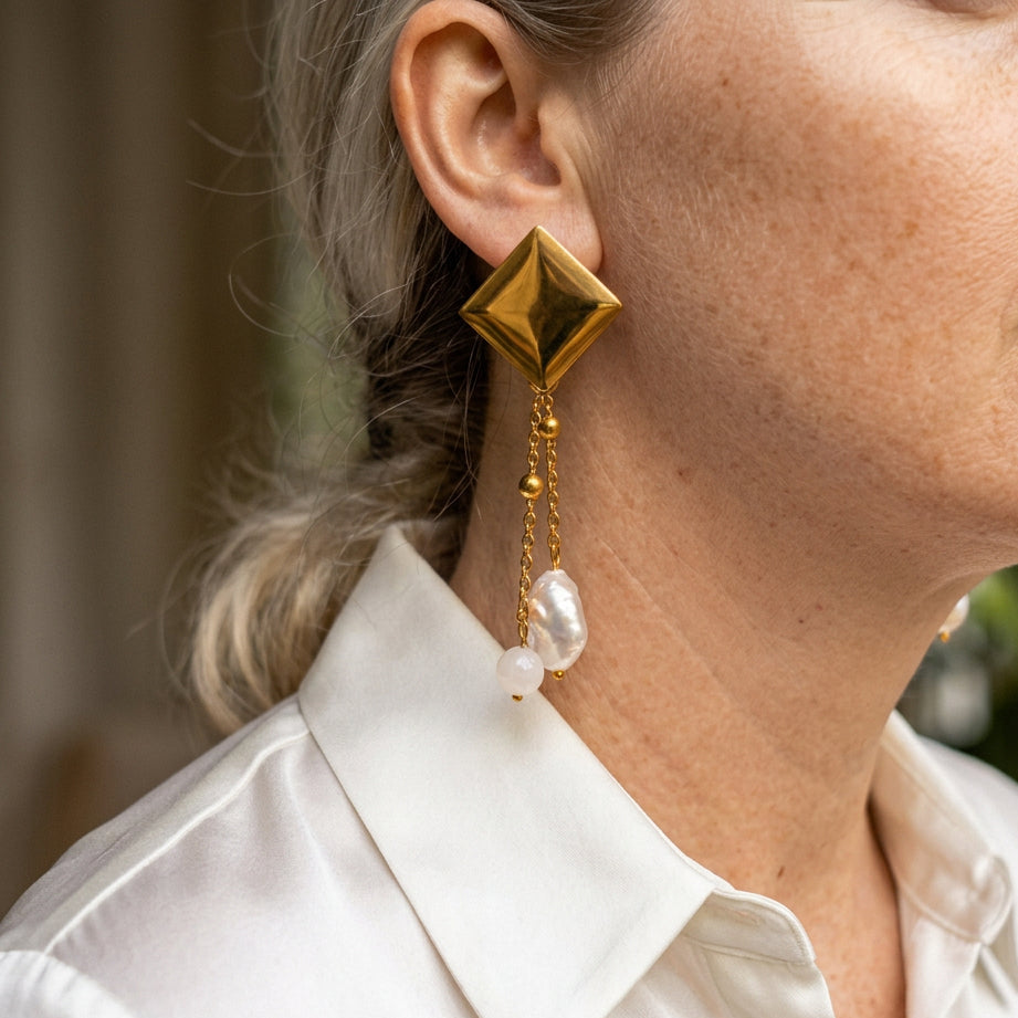 Gold-plated diamond-shaped stud earring with two fine beaded chains ending separately in a white oval baroque pearl and pale pink quartz bead, worn above a white shirt collar.