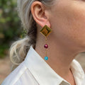 Gold-plated pyramid stud earring with a fine beaded chain, burgundy baroque pearl and bright blue topaz drop, worn above a white shirt collar.