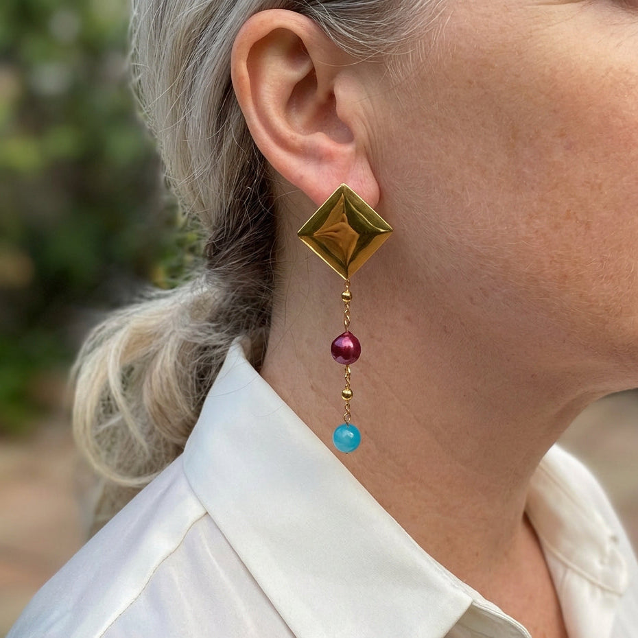 Gold-plated pyramid stud earring with a fine beaded chain, burgundy baroque pearl and bright blue topaz drop, worn above a white shirt collar.