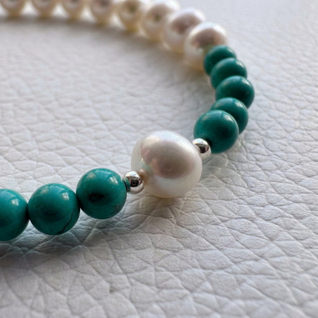 Close-up of a white baroque pearl framed by sterling silver beads between groups of Hubei turquoise beads on a white textured surface.