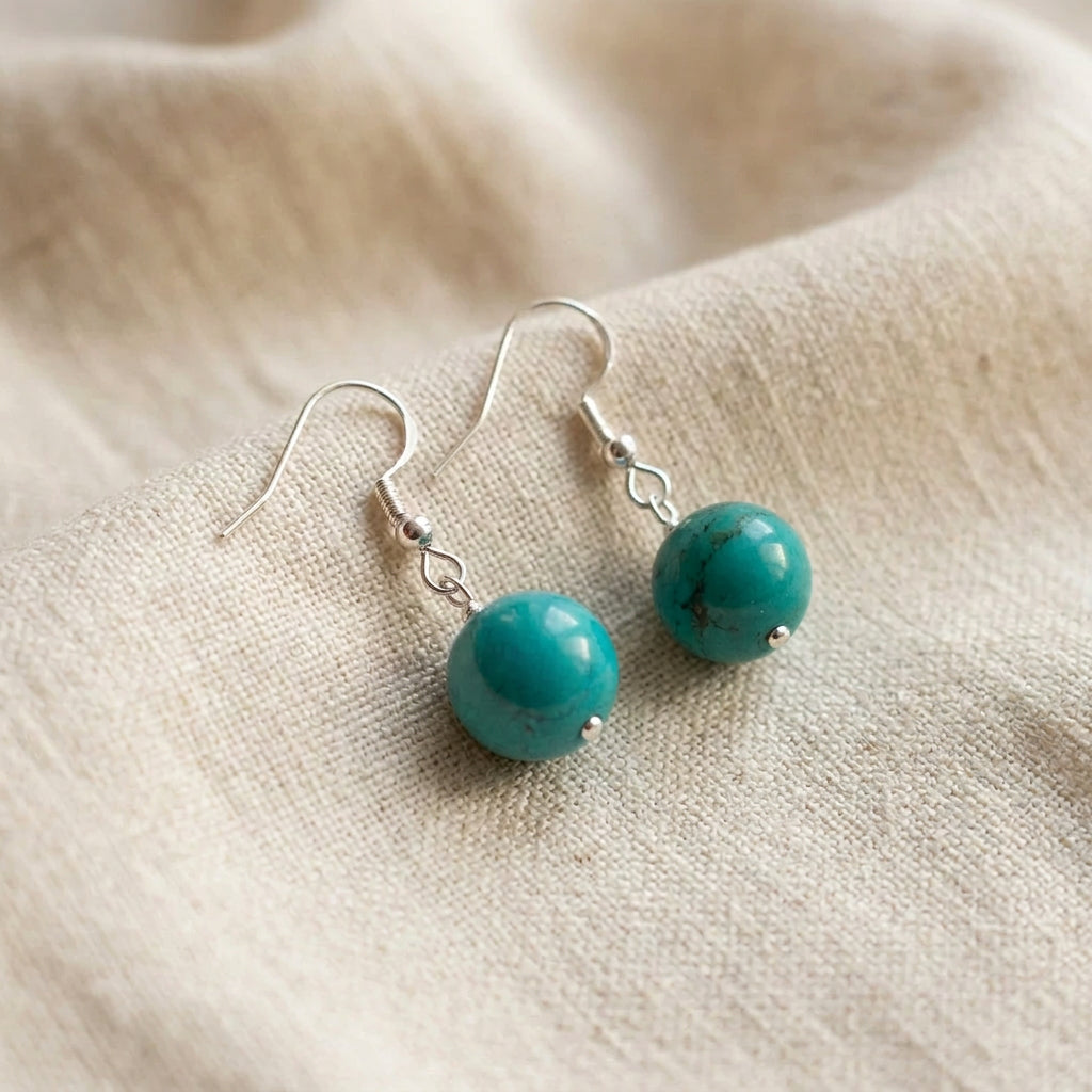 Angled close-up of Hubei turquoise earrings showing dark stone markings and sterling silver hook fittings on cream linen.