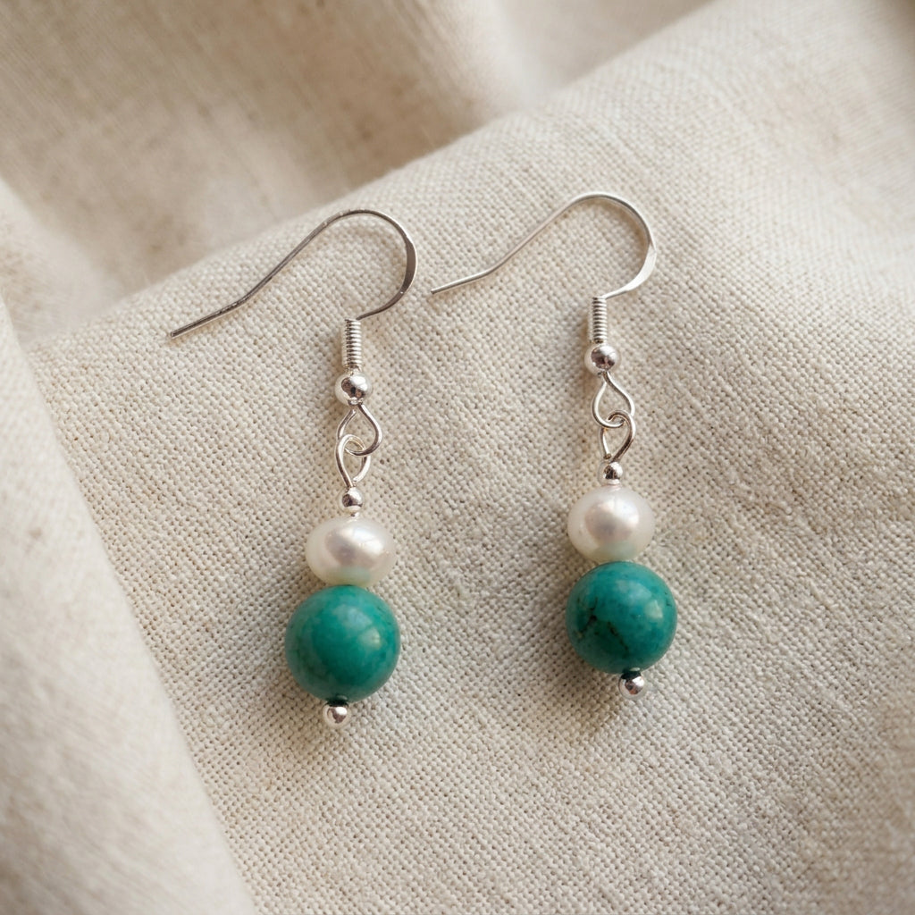 Pair of white baroque pearl and turquoise drop earrings with sterling silver hooks laid flat on a cream linen surface.