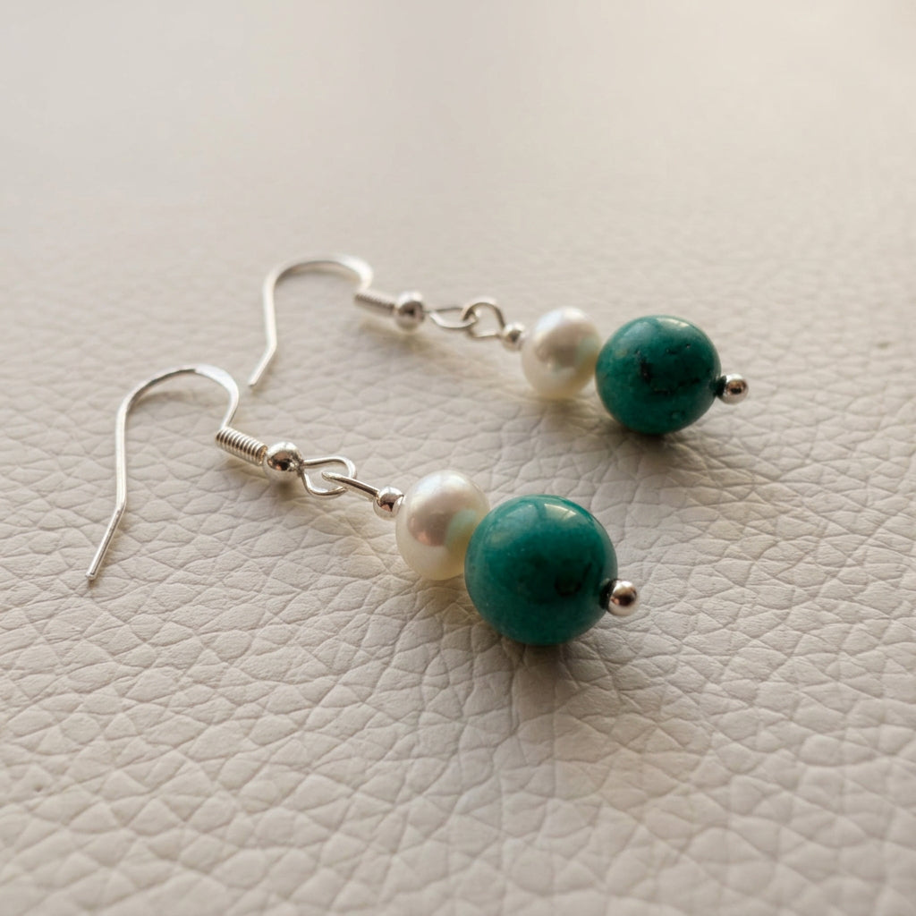 Angled view of white baroque pearl and turquoise drop earrings with sterling silver hooks resting on a white textured surface.
