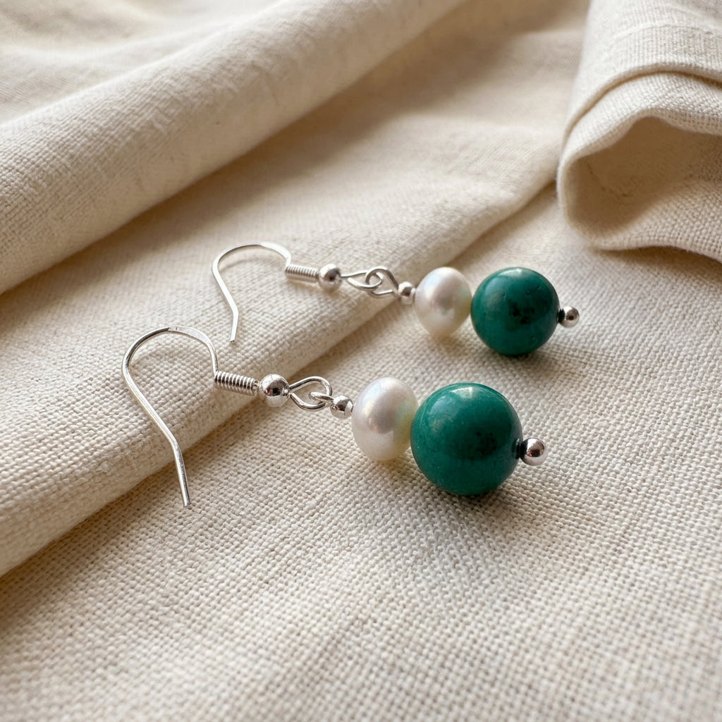 White baroque pearl and turquoise drop earrings with sterling silver hooks lying diagonally on softly folded cream linen.