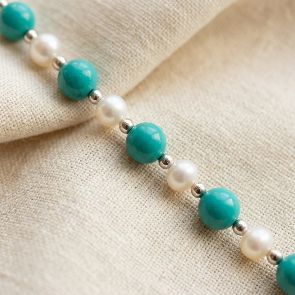 Hubei turquoise beads and white baroque pearls running diagonally across cream linen, separated by small silver-coloured spacers.