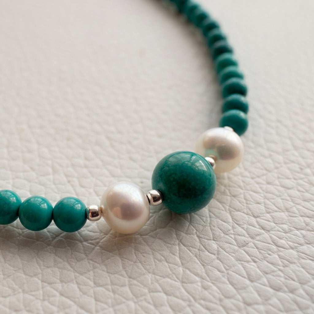 Close-up of the necklace’s central Hubei turquoise bead and two white baroque pearls on a white textured surface.