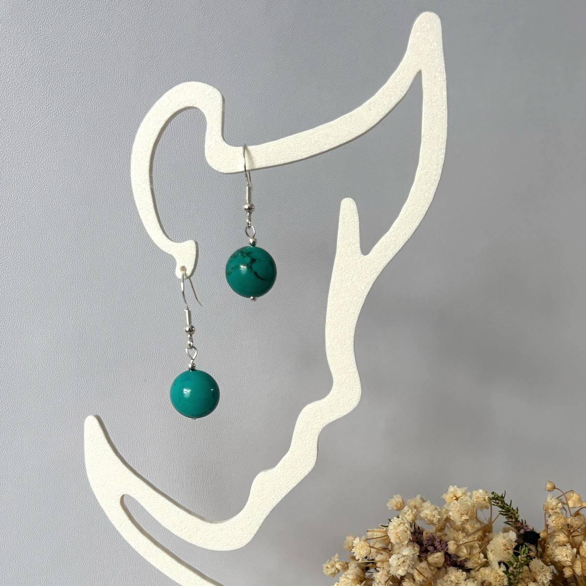 Sterling silver Hubei turquoise drop earrings hanging from a cream display stand beside dried flowers.